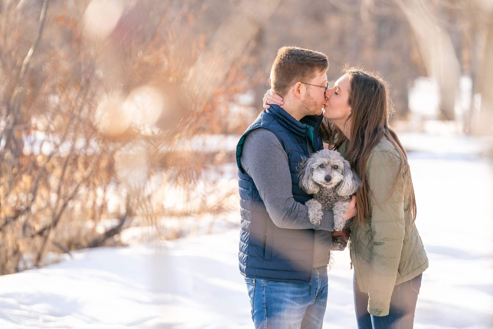 A man and woman kissing while cradling their miniature poodle between them on a winter day.